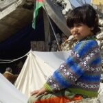 A young displaced child walking among tents and damaged buildings in Gaza.